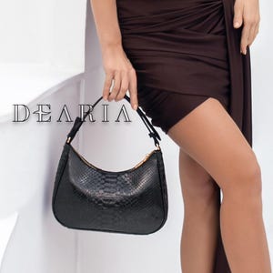 Könnte beinhalten: Ein braunes, ärmelloses Kleid mit drapiertem Rock und passender Handtasche. Die Handtasche hat ein Schlangenmuster. Das Kleid wird mit schwarzen Ballerinas und Goldschmuck kombiniert. Das Wort "DEARIA" ist auf dem Bild aufgedruckt.