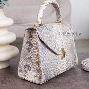K&ouml;nnte beinhalten: Eine strukturierte Handtasche mit Schlangenmuster in Grau- und Beiget&ouml;nen. Die Tasche hat einen oberen Griff, einen Klappverschluss mit goldfarbenem Verschluss und den Text "DEARIA". Ein passender Riemen ist sichtbar.