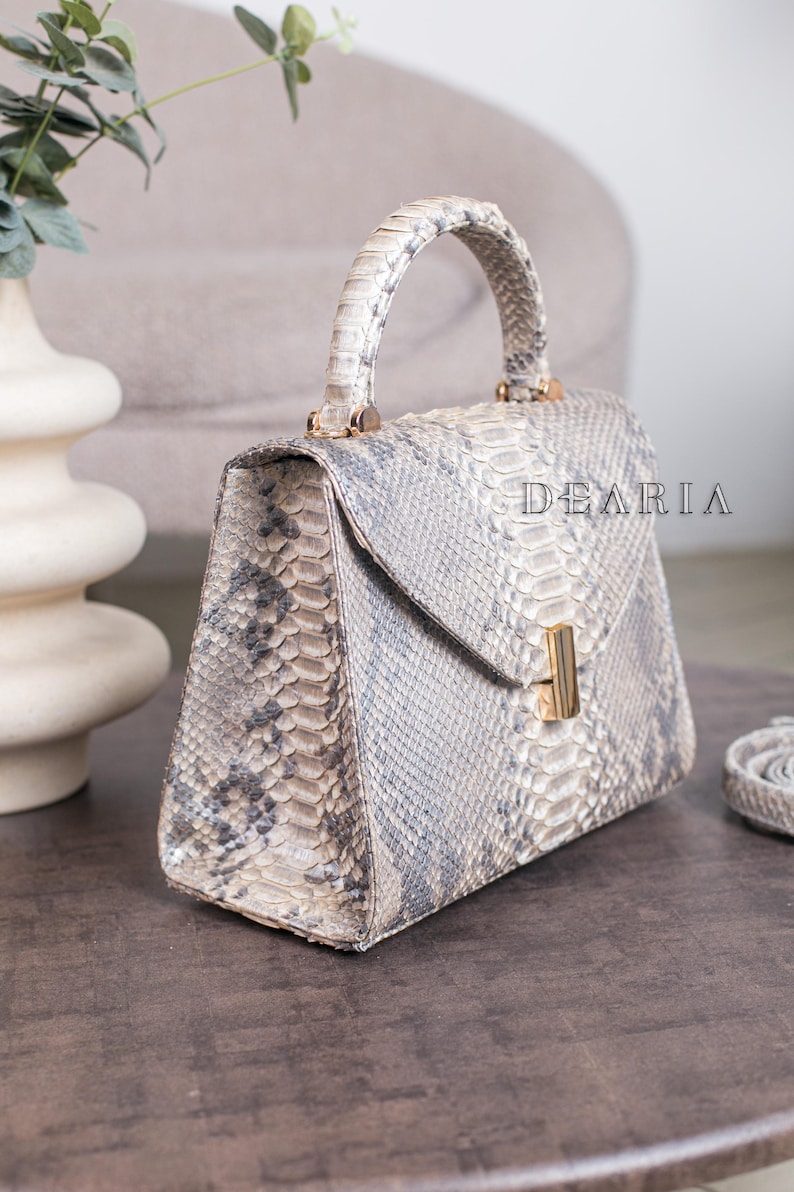 K&ouml;nnte beinhalten: Eine strukturierte Handtasche mit Schlangenmuster in Grau- und Beiget&ouml;nen. Die Tasche hat einen oberen Griff, einen Klappverschluss mit goldfarbenem Verschluss und den Text "DEARIA". Ein passender Riemen ist sichtbar.