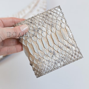 Könnte beinhalten: Ein quadratisches, silber-beige Schlangenleder-Portemonnaie. Die Brieftasche wird in einer Hand vor einem weißen Hintergrund gehalten. Das Wort "DEARIA" ist auf der Brieftasche aufgedruckt. Das Schlangenmuster ist sichtbar.