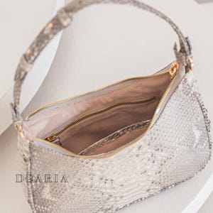 Pu&ograve; includere: Una borsa a mano in pelle di serpente grigio chiaro con una cerniera dorata e una tracolla a catena dorata. La borsa &egrave; aperta e l'interno &egrave; foderato con un tessuto beige chiaro.