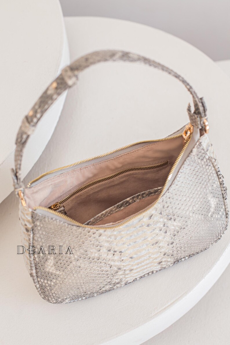 Pu&ograve; includere: Una borsa a mano in pelle di serpente grigio chiaro con una cerniera dorata e una tracolla a catena dorata. La borsa &egrave; aperta e l'interno &egrave; foderato con un tessuto beige chiaro.