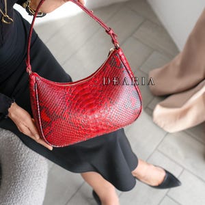 May include: Red snakeskin-effect shoulder bag with a curved design. The bag features a zip closure and a matching red strap. The word "DEARIA" is printed on the side. The bag is held by a person wearing a black outfit.