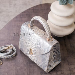 K&ouml;nnte beinhalten: Eine silber-graue Handtasche aus Schlangenleder mit einem oberen Griff und einem abnehmbaren Schulterriemen. Die Tasche hat eine rechteckige Form mit einem Klappenverschluss und goldfarbener Hardware. Das Wort "DEARIA" ist auf der Vorderseite der Tasche aufgedruckt.