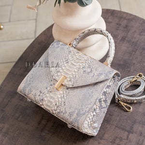 K&ouml;nnte beinhalten: Quadratische Handtasche mit Schlangenmuster in Grau- und Wei&szlig;t&ouml;nen. Sie hat einen oberen Griff, einen goldfarbenen Verschluss und einen abnehmbaren Schultergurt. Der Markenname "DEARIA" ist sichtbar.