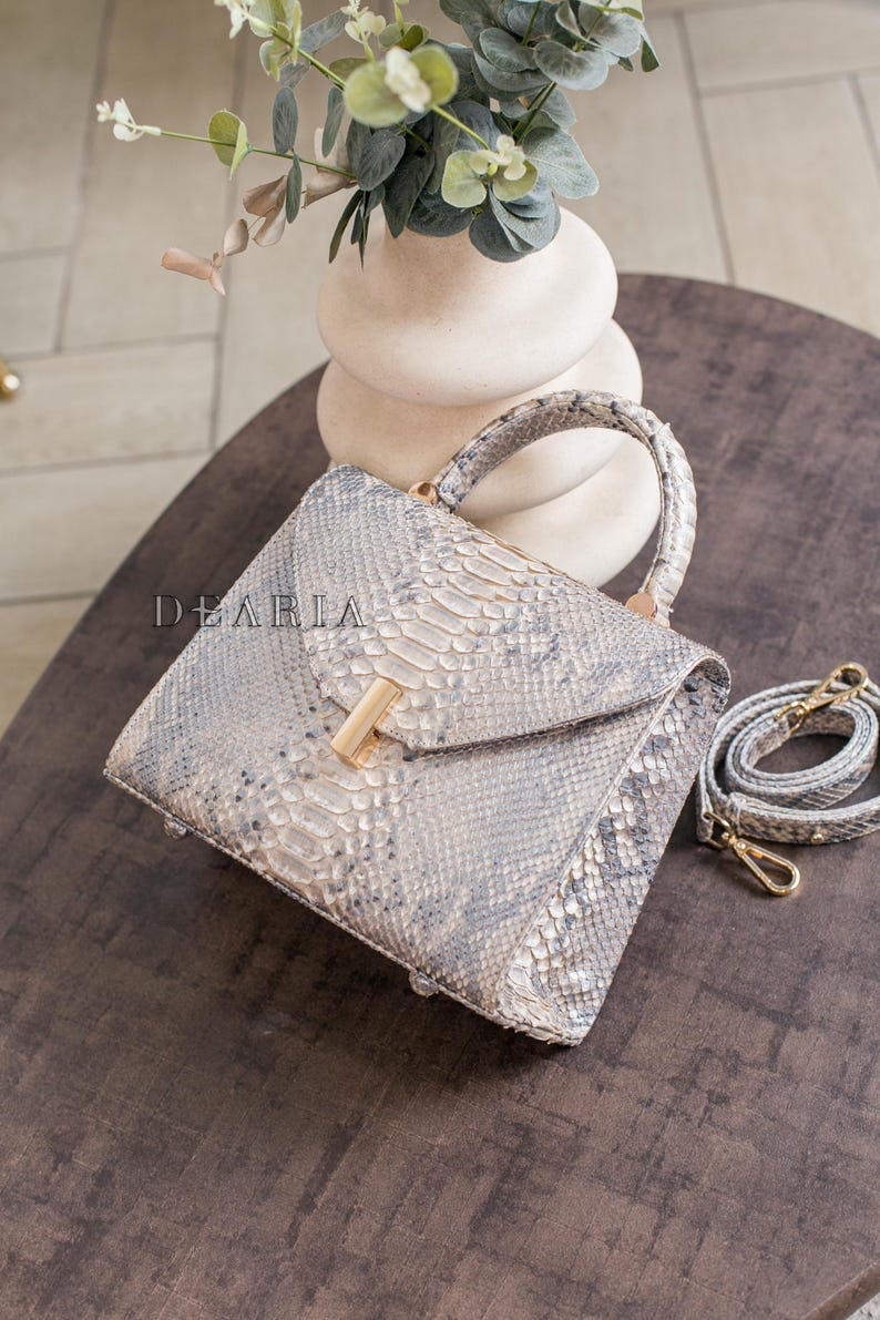 K&ouml;nnte beinhalten: Quadratische Handtasche mit Schlangenmuster in Grau- und Wei&szlig;t&ouml;nen. Sie hat einen oberen Griff, einen goldfarbenen Verschluss und einen abnehmbaren Schultergurt. Der Markenname "DEARIA" ist sichtbar.