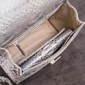 K&ouml;nnte beinhalten: Ge&ouml;ffnete Handtasche mit Schlangenmuster in Grau- und Beiget&ouml;nen, mit passender Clutch im Inneren. Die Tasche hat einen goldfarbenen Verschluss und ein hellbraunes Innenfutter. Der Markenname "DEARIA" ist sichtbar.