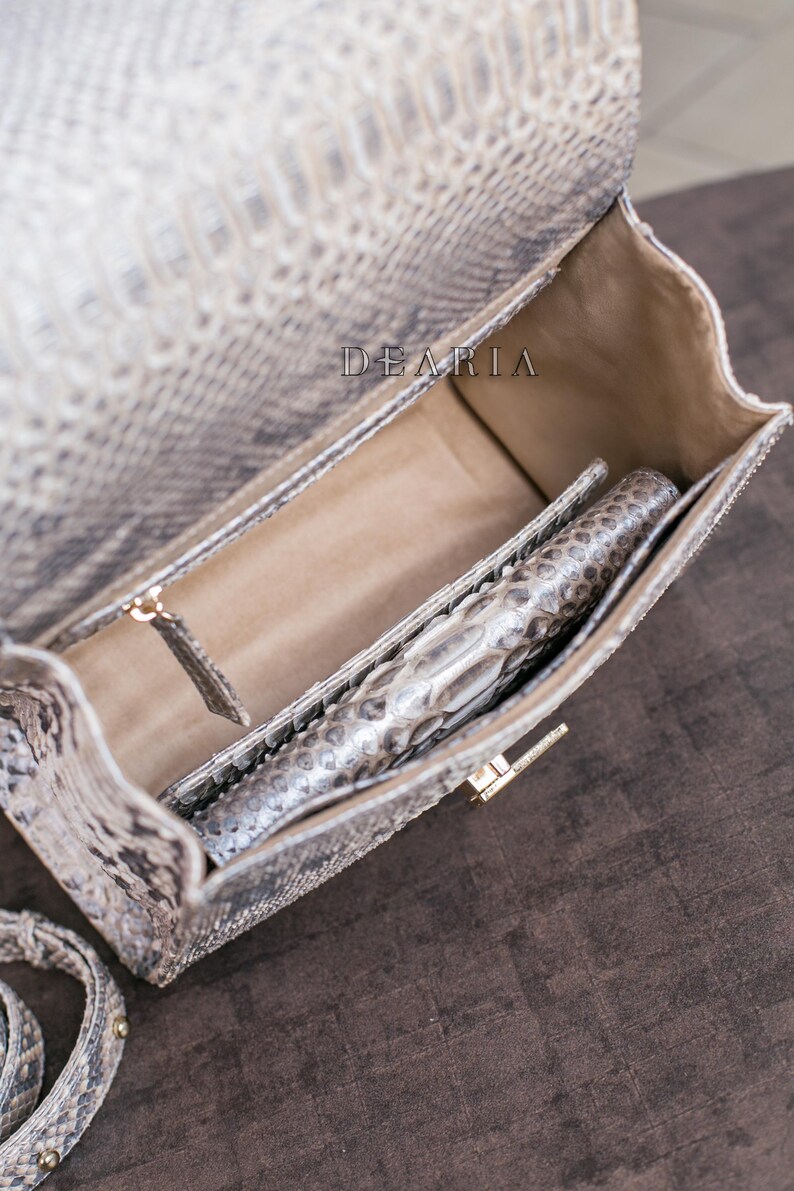 K&ouml;nnte beinhalten: Ge&ouml;ffnete Handtasche mit Schlangenmuster in Grau- und Beiget&ouml;nen, mit passender Clutch im Inneren. Die Tasche hat einen goldfarbenen Verschluss und ein hellbraunes Innenfutter. Der Markenname "DEARIA" ist sichtbar.