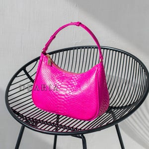 May include: A bright pink snakeskin-textured handbag with a gold zipper and a single top handle. The bag is sitting on a black metal chair.