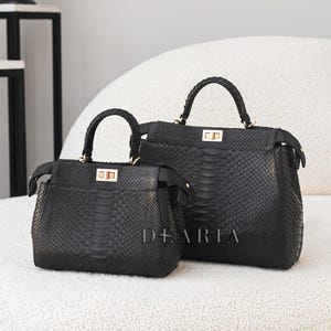 May include: Two black handbags with a snakeskin texture. The bags have top handles and gold-coloured clasps. The smaller bag is in front of the larger one. The word "DEARIA" is printed on the front of the bags.
