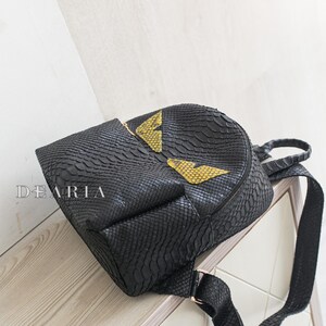 May include: Black backpack with a snakeskin texture. It has a gold zip and two yellow and gold eye-shaped designs. The brand name "DEARIA" is printed on the front.