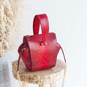 May include: A red handbag with a snakeskin pattern, featuring a top handle and a gold chain strap. The bag is sitting on a wooden stool. The word "DEARIA" is printed on the side of the bag.