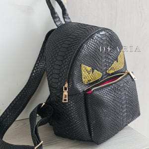 May include: Black snakeskin-textured backpack with gold-coloured zips and a front pocket. It has yellow and black eye-shaped details. The word "DEARIA" is printed on the side.