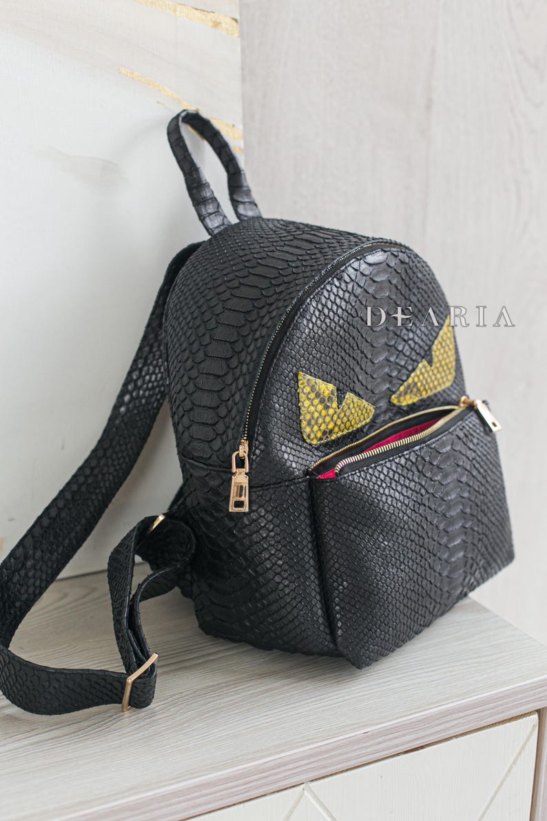 May include: Black snakeskin-textured backpack with gold-coloured zips and a front pocket. It has yellow and black eye-shaped details. The word "DEARIA" is printed on the side.