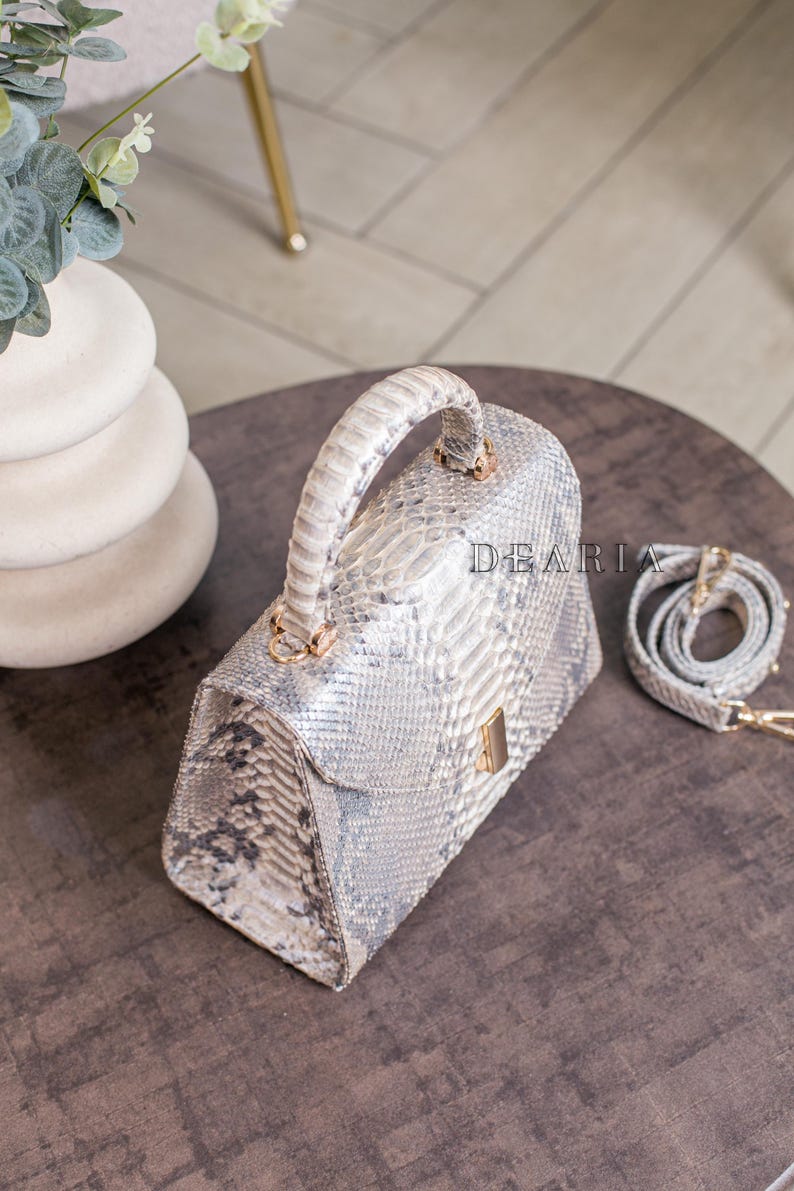 K&ouml;nnte beinhalten: Eine hellgraue Handtasche mit Schlangenmuster, einem oberen Griff und einem abnehmbaren Schulterriemen. Die Tasche hat goldfarbene Hardware und eine rechteckige Form. Das Wort "DEARIA" ist auf der Tasche aufgedruckt.