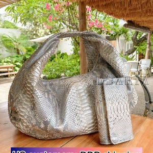 May include: A silver-toned, textured handbag with a matching wallet. The bag has a curved shape and a single handle. The wallet is rectangular. The products are made of a material with a scale-like pattern.