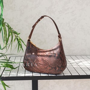 Pu&ograve; includere: Una borsa a mano marrone con motivo a pelle di serpente e una tracolla corta. La borsa &egrave; appoggiata su una superficie piastrellata bianca.