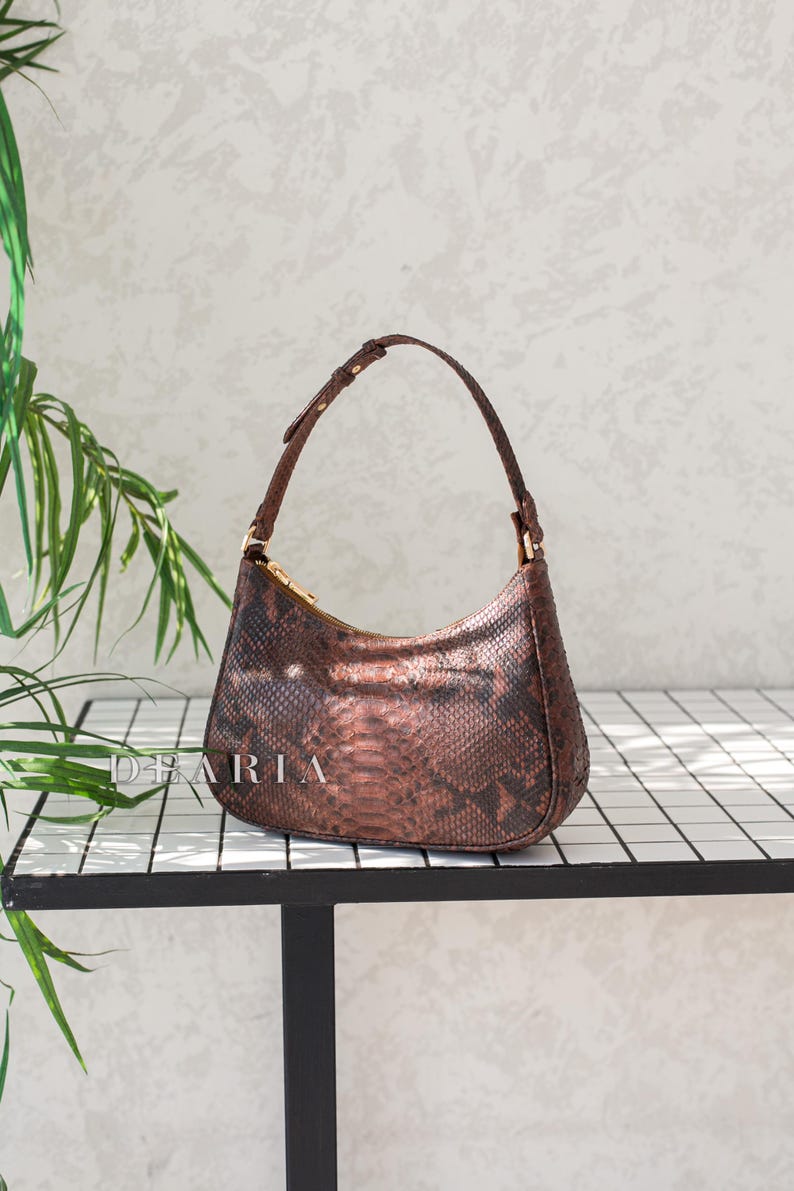 Pu&ograve; includere: Una borsa a mano marrone con motivo a pelle di serpente e una tracolla corta. La borsa &egrave; appoggiata su una superficie piastrellata bianca.
