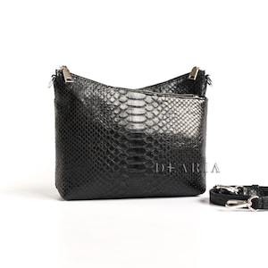 May include: Black leather crossbody bag with a snakeskin pattern. The bag has a rectangular shape with a zippered top and a detachable shoulder strap. The brand name "DEARIA" is printed on the front.