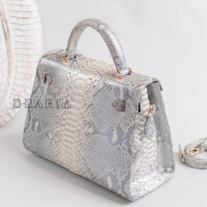 May include: A silver snakeskin-patterned handbag with a top handle and a detachable shoulder strap. The bag has a rectangular shape with a flap closure and gold-coloured hardware. The word "DEARIA" is printed on the front.