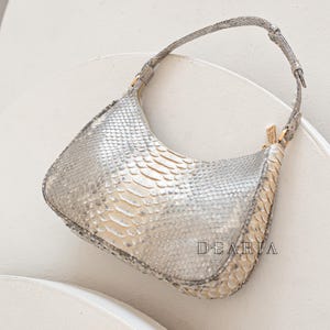 Pu&ograve; includere: Una borsa a mano in pelle di serpente argentata con un cinturino corto per la spalla. La borsa ha una chiusura a zip e una forma leggermente curva.