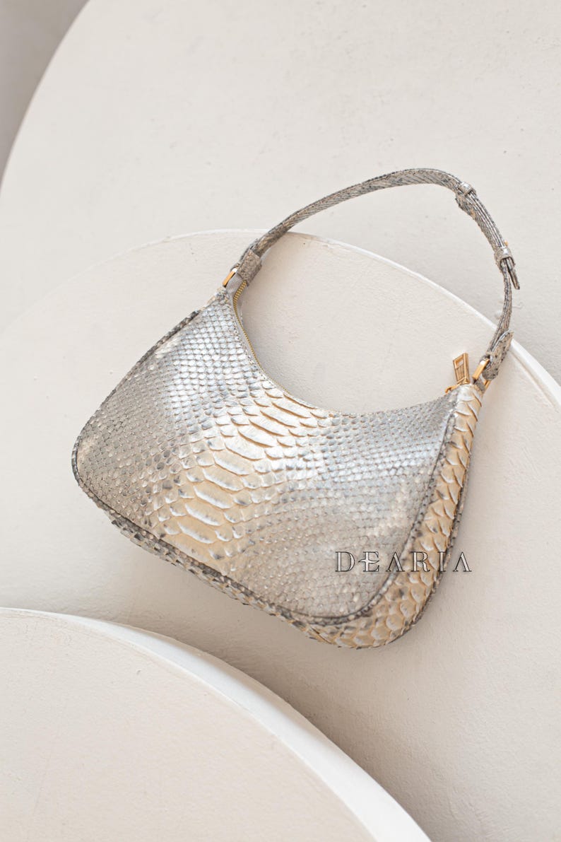 Pu&ograve; includere: Una borsa a mano in pelle di serpente argentata con un cinturino corto per la spalla. La borsa ha una chiusura a zip e una forma leggermente curva.