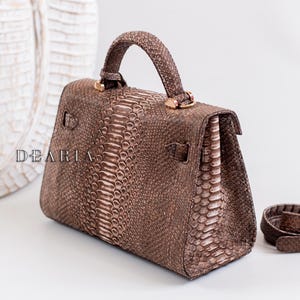 May include: A brown handbag with a snakeskin texture. The bag features a top handle, a flap closure, and gold-coloured hardware. A matching shoulder strap is also visible. The word "DEARIA" is printed on the side of the bag.