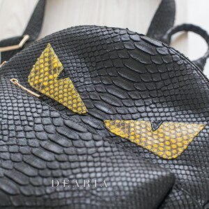 May include: Black leather backpack with a snakeskin texture. It has yellow and black lightning bolt-shaped details. Includes a top handle and shoulder straps. The brand name "DEARIA" is visible on the front.