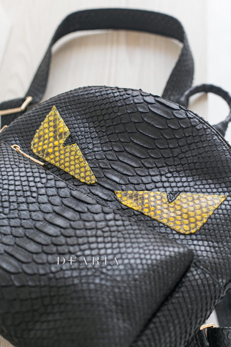 May include: Black leather backpack with a snakeskin texture. It has yellow and black lightning bolt-shaped details. Includes a top handle and shoulder straps. The brand name "DEARIA" is visible on the front.