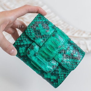 Op de afbeelding: Een groene en zwarte clutch met slangenpatroon en klepsluiting. De clutch heeft een rechthoekige vorm met een textuur. Het woord "DEARIA" is linksboven zichtbaar.
