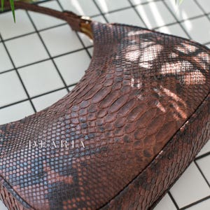Pu&ograve; includere: Una borsa a mano in pelle di serpente marrone con un manico corto. La borsa ha una superficie testurizzata e la parola "DEARLA" &egrave; stampata sul davanti.
