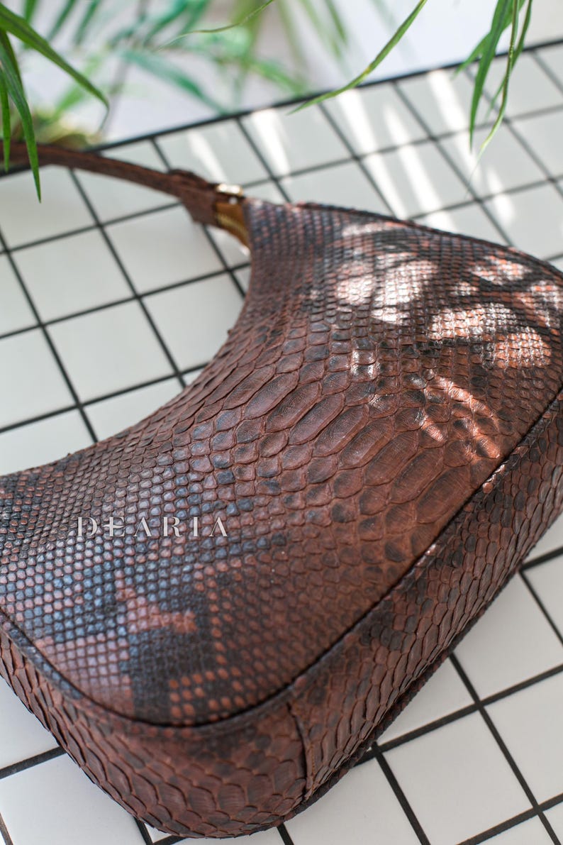 Pu&ograve; includere: Una borsa a mano in pelle di serpente marrone con un manico corto. La borsa ha una superficie testurizzata e la parola "DEARLA" &egrave; stampata sul davanti.