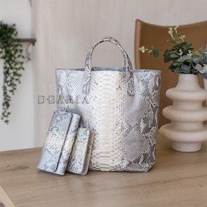 Peut inclure: Ensemble d'accessoires assortis, comprenant un sac fourre-tout et deux portefeuilles, tous avec un motif peau de serpent gris et blanc. Le sac fourre-tout a une poignée et la marque "DEARIA" est visible.