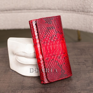Może przedstawiać: Czerwony skórzany portfel z wzorem skóry węża. Portfel jest prostokątny z zapięciem na klapę. Słowo "DEARIA" jest wydrukowane na biało z przodu. Portfel jest prezentowany obok białej rzeźby.
