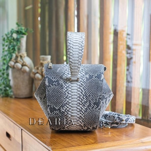 May include: A gray and white snakeskin-patterned handbag with a structured design and a top handle. The bag features a zipper closure and a detachable strap. The word "DEARIA" is printed on the front of the bag.