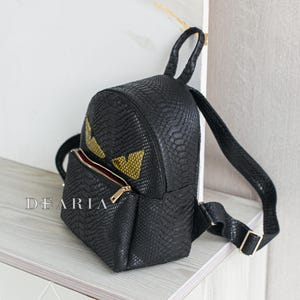 May include: Black faux leather backpack with a reptile skin texture. The backpack features a gold zip, a small front pocket, and yellow eye-shaped accents. The word "DEARIA" is printed on the front.
