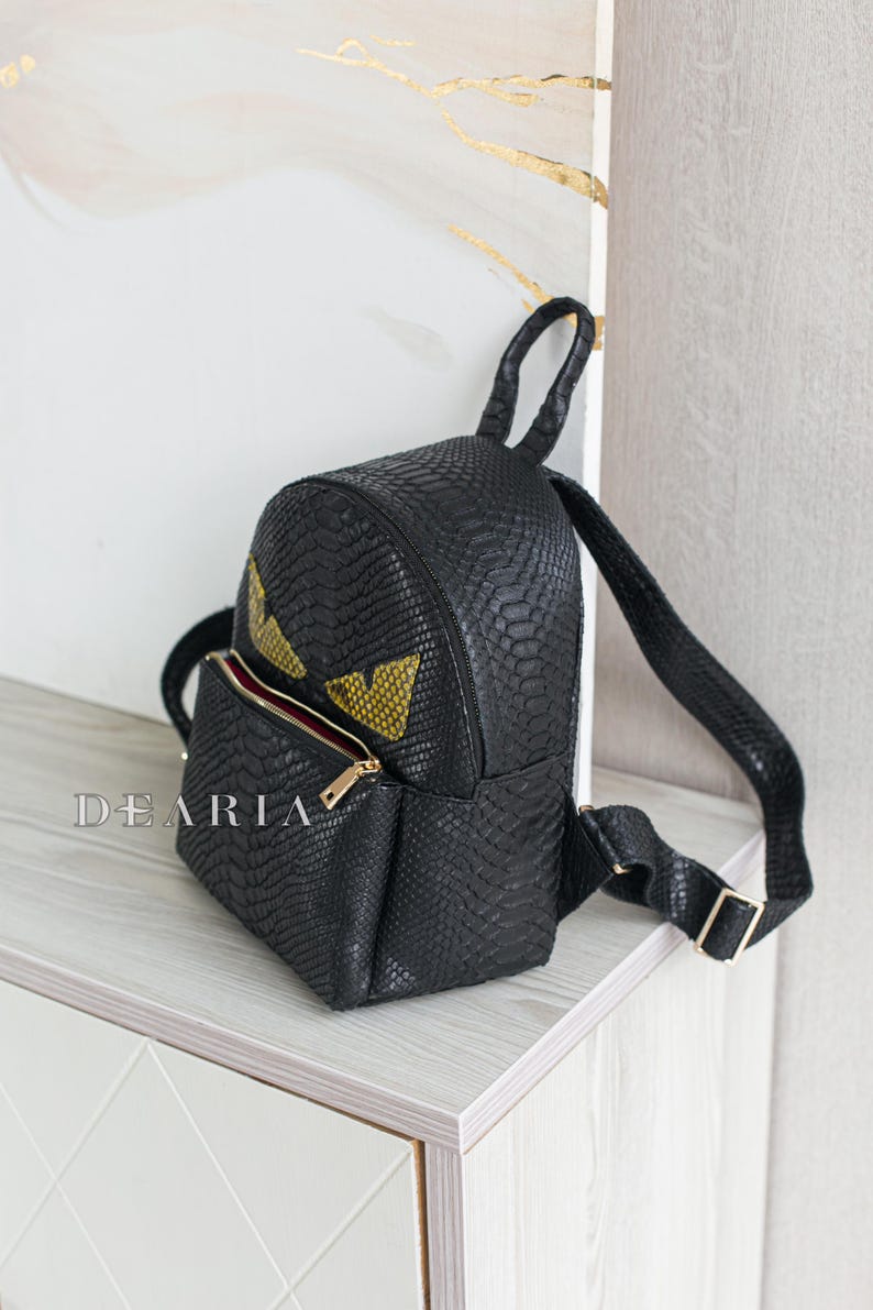 May include: Black faux leather backpack with a reptile skin texture. The backpack features a gold zip, a small front pocket, and yellow eye-shaped accents. The word "DEARIA" is printed on the front.