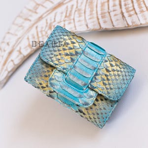 Pode incluir: Uma clutch azul e dourada de pele de cobra com fecho de aba e alça a condizer. A palavra "DEARIA" está impressa na frente da clutch. A clutch é feita de um material escamoso.
