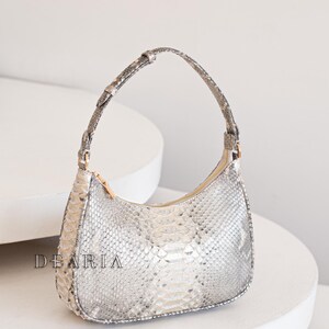 Pu&ograve; includere: Una piccola borsa a mano in pelle di serpente grigio chiaro con un manico corto e curvo. La borsa ha una chiusura a zip e un motivo testurizzato sottile.