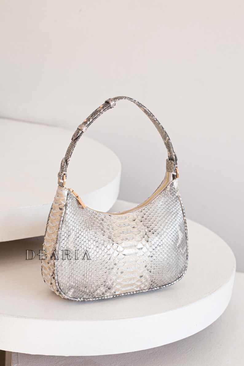 Pu&ograve; includere: Una piccola borsa a mano in pelle di serpente grigio chiaro con un manico corto e curvo. La borsa ha una chiusura a zip e un motivo testurizzato sottile.