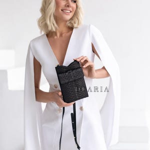 Könnte beinhalten: Eine schwarze Crossbody-Tasche aus strukturiertem Leder mit Klappe und langem Riemen. Die Tasche wird von einer Person gehalten, die ein weißes Kleid mit goldenen Knöpfen trägt. Der Text "DEARIA" ist auf der Tasche zu sehen.