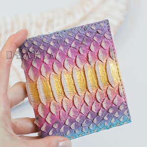 May include: A vibrant wallet featuring a scale design in hues of purple, pink, blue, and gold. The word "DEARIA" is printed on the wallet. The wallet is held by a person.