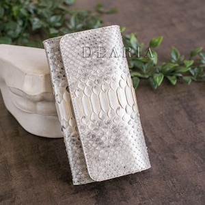 May include: A silver and white snakeskin-textured wallet with the word "DEARIA" printed on the front. The wallet is closed and has a flap closure. The background is a dark brown surface with green foliage.