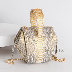 May include: A small, grey and gold snakeskin handbag with a gold chain strap and a gold snakeskin top handle. The bag has a geometric shape and the brand name "DEARIA" is visible on the side.