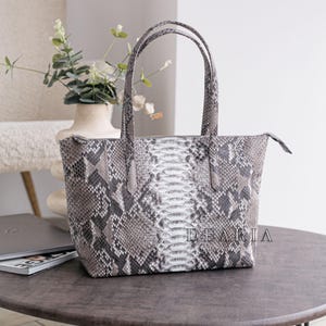 May include: A gray and white snakeskin-patterned tote bag with two handles. The bag has a zipper closure and the word "DEARIA" is printed on the front. The bag is sitting on a dark brown table.