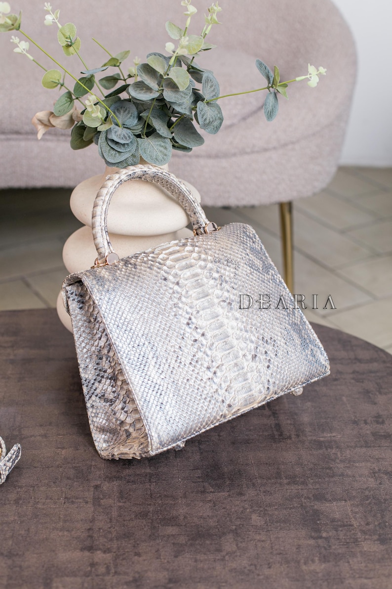 K&ouml;nnte beinhalten: Eine silber-graue Handtasche mit Schlangenmuster. Die Tasche hat einen oberen Griff und eine rechteckige Form. Das Wort "DEARIA" ist auf der Seite aufgedruckt. Die Tasche liegt auf einer dunkelbraunen Oberfl&auml;che.