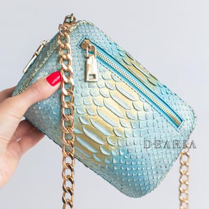 Könnte beinhalten: Rechteckige, hellblaue Handtasche mit goldener Kette und Reißverschluss. Die Tasche hat ein Schlangenmuster mit goldenen Akzenten. Reißverschluss und Kette sind goldfarben. Der Hintergrund ist weiß.