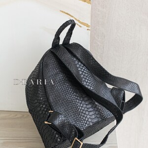 May include: Black backpack with a textured, snake-like pattern. The bag has adjustable straps, a top handle, and gold-coloured hardware. The word "DEARIA" is printed on the front of the bag.
