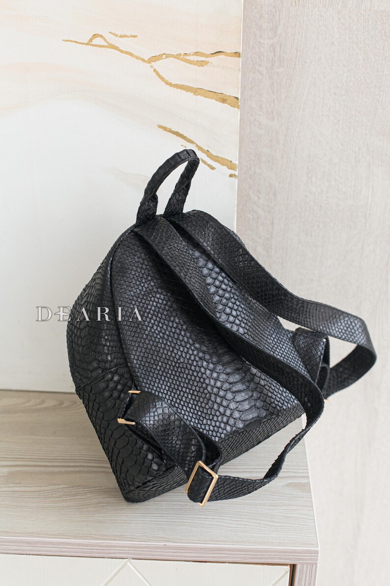 May include: Black backpack with a textured, snake-like pattern. The bag has adjustable straps, a top handle, and gold-coloured hardware. The word "DEARIA" is printed on the front of the bag.