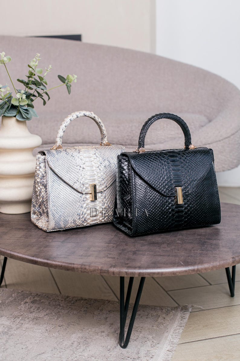 K&ouml;nnte beinhalten: Zwei Lederhandtaschen, eine in Schwarz und die andere in einem hellgrauen Schlangenmuster, stehen auf einem runden Tisch. Beide Taschen haben eine strukturierte Form, obere Griffe und goldfarbene Beschl&auml;ge. Die Taschen sind mit dem Text "DEMIA" beschriftet.